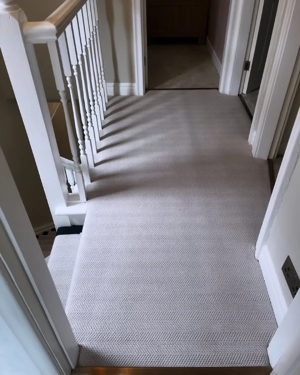 Light beige herringbone patterned carpet fitted in a hallway with white railings and doors leading to rooms.