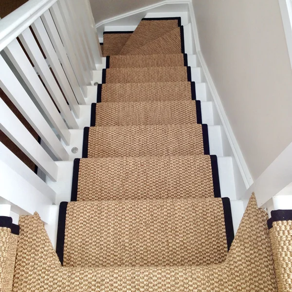 An overhead view looking down a white wooden staircase featuring a textured, tan sisal carpet runner with a bold black fabric border.
