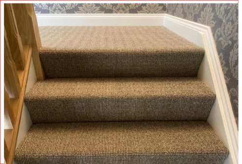 Close-up of a staircase featuring a textured brown loop carpet with white baseboards and patterned wallpaper.