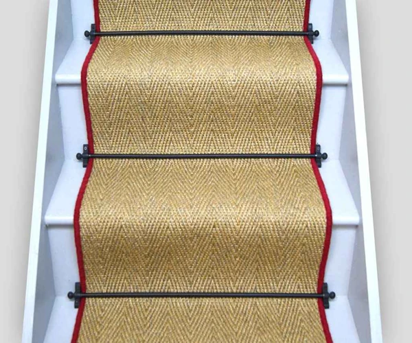 Natural tan chevron sisal stair runner with a red border and black metal rods on white stairs.