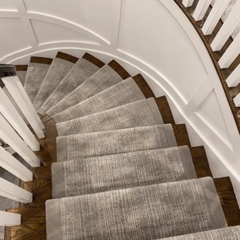 curved wooden staircase with a gray textured carpet runner and white wainscoted walls.