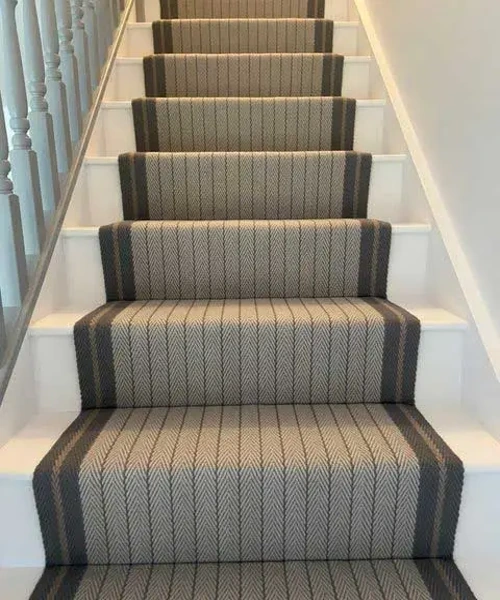narrow wool carpet runner with a grey and beige herringbone pattern and dark grey borders, installed on a white wooden staircase.