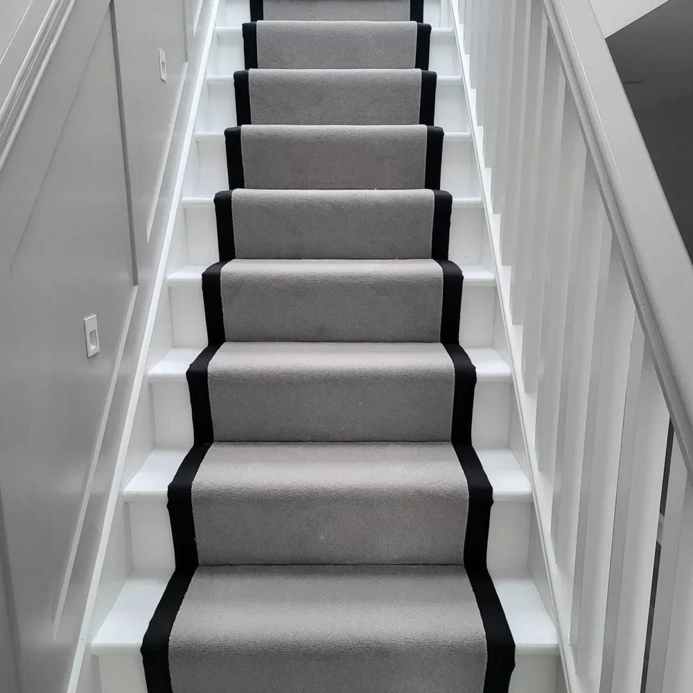 straight white staircase featuring a plush light gray wool carpet runner with a bold black fabric border