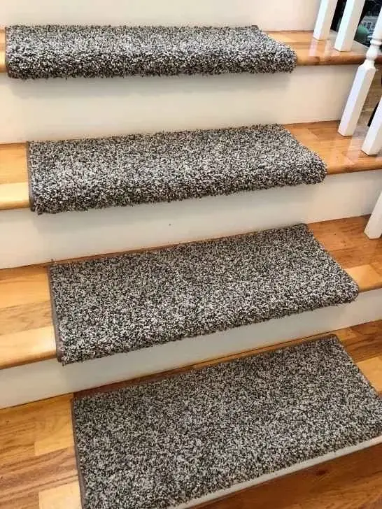 A close-up, angled view of a light oak staircase with white risers, fitted with rectangular, speckled gray and brown plush carpet treads.