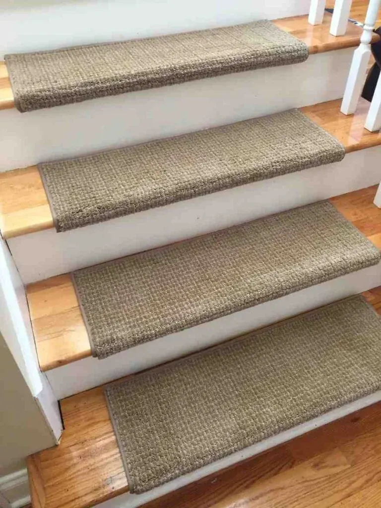 An angled, close-up view of a wooden staircase featuring rectangular, woven beige carpet treads on light oak steps with white risers.