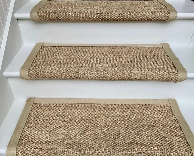 Woven sisal stair treads with tan fabric borders on white painted wooden steps.